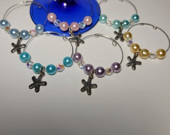 May include: Set of six wine glass charms with silver-tone hoops. Each charm features a different color of faux pearls, iridescent beads, and a starfish charm. Colors include light blue, pink, teal, lavender, and gold.