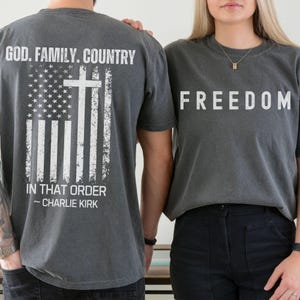 May include: Two charcoal gray t-shirts. One displays "GOD. FAMILY. COUNTRY" with an American flag and cross design, and the text "IN THAT ORDER ~ CHARLIE KIRK." The other shirt has the word "FREEDOM" in white block letters.
