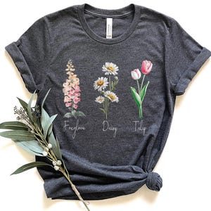 May include: Dark gray t-shirt featuring watercolor illustrations of a foxglove, daisies, and a tulip. The names of the flowers are written below each image. The shirt is folded at the sleeves and knotted at the bottom.
