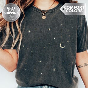 May include: Dark gray t-shirt with a celestial design featuring stars and a crescent moon. The shirt has the word "Pepper" at the bottom. The shirt also has the words "Next Day Shipping" and "Comfort Colors" on it.