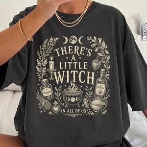 May include: A charcoal gray t-shirt featuring a cream-colored graphic print with the text "THERE'S A LITTLE WITCH IN ALL OF US." The design includes illustrations of a cauldron, potion bottles, a candle, herbs, and moon phases.