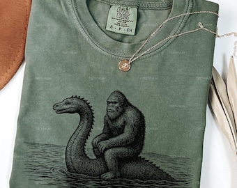 May include: Moss green Comfort Colors t-shirt featuring a black and white illustration of a gorilla riding a sea serpent. The shirt has a crew neck and short sleeves. A gold necklace with a pendant is draped on the shirt.
