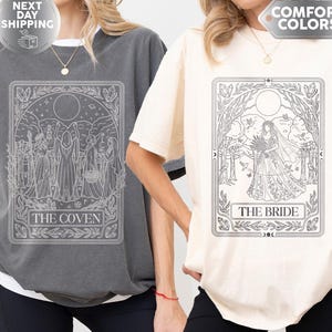 May include: A charcoal gray t-shirt featuring a white line art graphic labeled THE COVEN and an ivory t-shirt featuring a white line art graphic labeled THE BRIDE. Both shirts have short sleeves, a crew neckline, and a relaxed, loose-fitting silhouette.