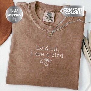 May include: Espresso-colored t-shirt with the text "hold on, I see a bird" and a graphic of binoculars. The shirt has a crew neck and a tag that says "Comfort Colors". A silver necklace with a pendant is also visible.