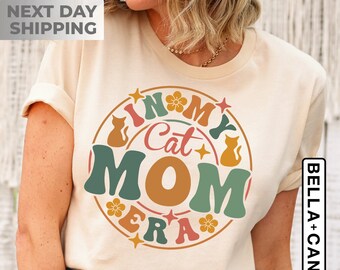 May include: A beige t-shirt with a retro-style graphic design that says "In My Cat Mom Era" in a circle with cats and flowers. The design is in shades of green, orange, and yellow.