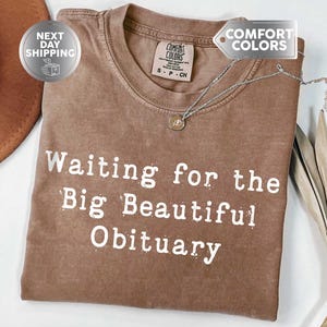 May include: Espresso-colored t-shirt with the text "Waiting for the Big Beautiful Obituary" in white. The shirt has a crew neck and a Comfort Colors tag. A silver necklace with a small pendant is draped on the shirt.