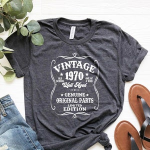 May include: Dark gray t-shirt with the text "VINTAGE 1970" in a white decorative frame. The shirt is paired with blue jeans and black sandals. The shirt's design includes the phrases "Old School", "Well Aged", and "Genuine Original Parts".