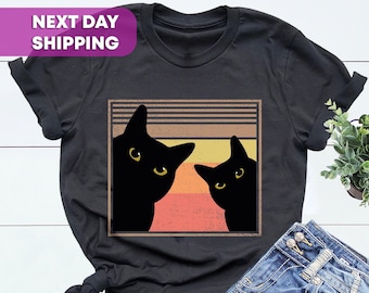 May include: Black t-shirt with a vintage-style graphic of two black cats peeking out from behind a striped window. The graphic has a faded, retro look with orange, yellow, and brown colors.