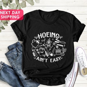 K&ouml;nnte beinhalten: Schwarzes T-Shirt mit wei&szlig;em Text "HOEING AIN'T EASY" und einer Grafik mit Gartenger&auml;ten und Pflanzen.