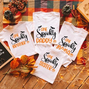 May include: White t-shirts with Halloween-themed graphics. Each shirt has the words "One Spooky" followed by "Brother," "Daddy," "Mama," and "Sister" in black and orange. Bats, spiderwebs, and pumpkins are also featured.
