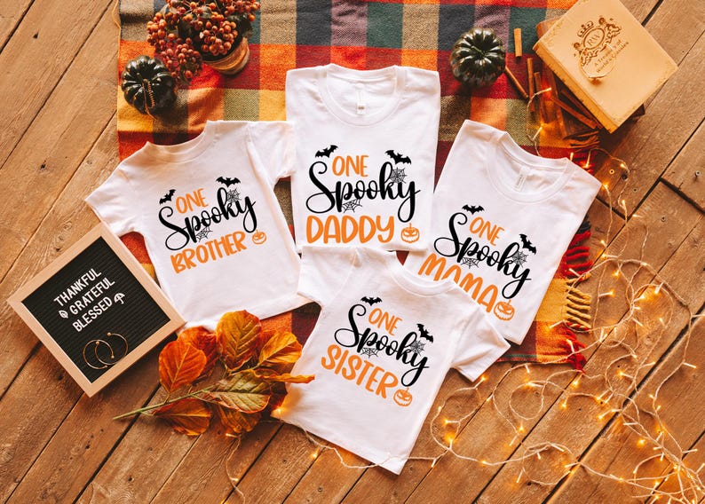 May include: White t-shirts with Halloween-themed graphics. Each shirt has the words "One Spooky" followed by "Brother," "Daddy," "Mama," and "Sister" in black and orange. Bats, spiderwebs, and pumpkins are also featured.