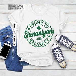 Op de afbeelding: Wit T-shirt met een groene cirkelvormige afbeelding met de tekst "*PRONE TO SHENANIGANS AND MALARKEY*" met een klavertje vier in de cirkel.