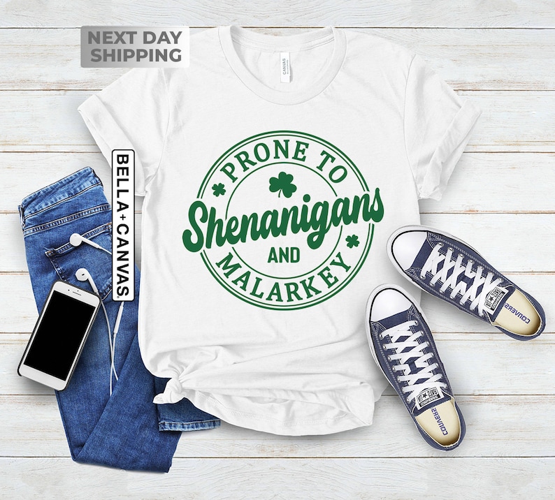 Op de afbeelding: Wit T-shirt met een groene cirkelvormige afbeelding met de tekst "*PRONE TO SHENANIGANS AND MALARKEY*" met een klavertje vier in de cirkel.