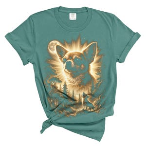 May include: Teal-colored t-shirt featuring a detailed illustration of a Chihuahua dog with a radiant halo, set against a forest and moon backdrop. The design includes a smaller dog silhouette. A unique graphic tee.
