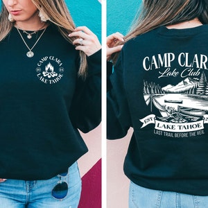 May include: Black sweatshirt with a white graphic on the front that says "Camp Clara Lake Tahoe" and a white graphic on the back that says "Camp Clara Lake Club" with a scene of a lake, mountains, and trees. The text on the back also says "Est. 2024" and "Last Trail Before The Veil."