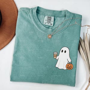 May include: Light green Comfort Colors t-shirt featuring a ghost graphic holding a coffee cup and a jack-o'-lantern. A gold necklace and a brown hat are also visible. The shirt is labelled "LIGHT GREEN".