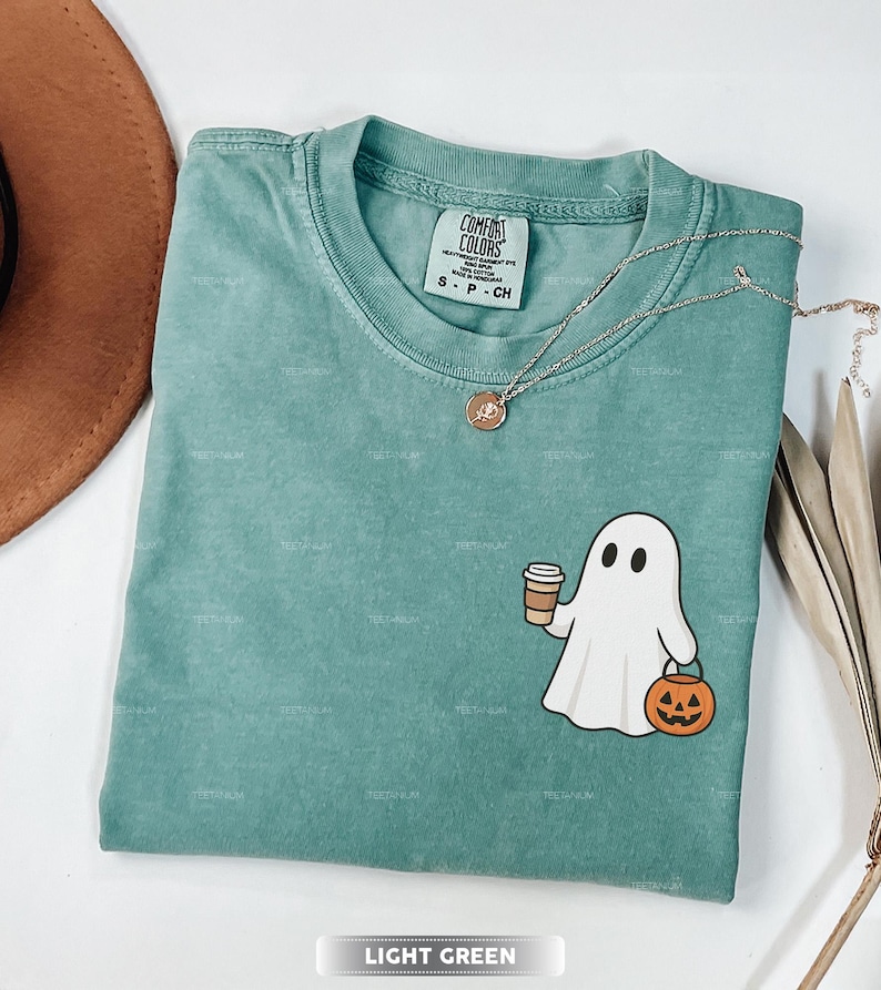 May include: Light green Comfort Colors t-shirt featuring a ghost graphic holding a coffee cup and a jack-o'-lantern. A gold necklace and a brown hat are also visible. The shirt is labelled "LIGHT GREEN".