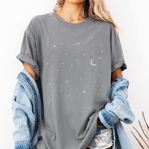 May include: Gray t-shirt with a celestial design featuring stars and a crescent moon. The shirt has a relaxed fit and rolled-up sleeves. The word "GRAY" is displayed in a banner at the bottom of the image.