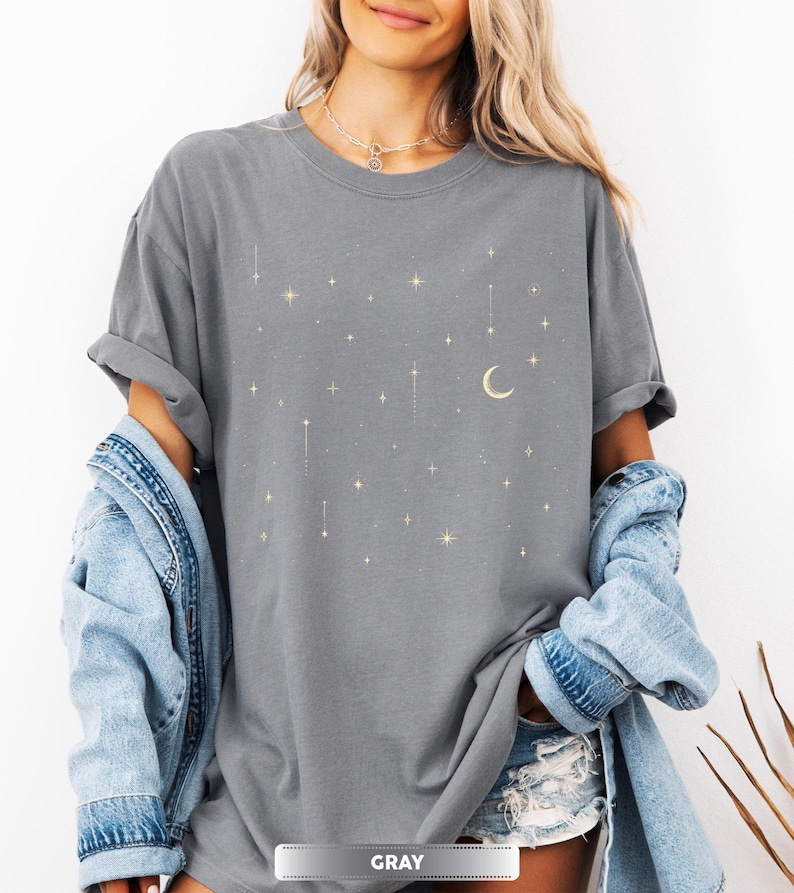 May include: Gray t-shirt with a celestial design featuring stars and a crescent moon. The shirt has a relaxed fit and rolled-up sleeves. The word "GRAY" is displayed in a banner at the bottom of the image.
