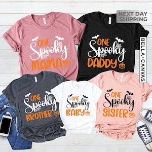 May include: A collection of five short-sleeved t-shirts in various colors, each featuring the words "One Spooky" followed by "Mama", "Daddy", "Brother", "Baby", and "Sister". The shirts have Halloween-themed graphics.