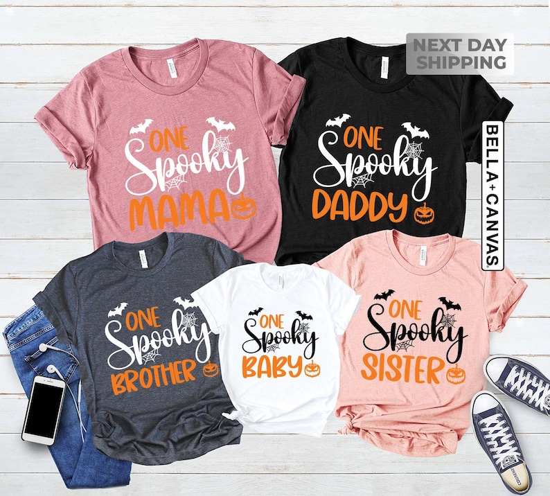 May include: A collection of five short-sleeved t-shirts in various colors, each featuring the words "One Spooky" followed by "Mama", "Daddy", "Brother", "Baby", and "Sister". The shirts have Halloween-themed graphics.