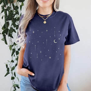 May include: Navy blue t-shirt with a celestial design featuring gold stars and a crescent moon. The word "MIDNIGHT" is printed at the bottom. The shirt has a crew neck and short sleeves.