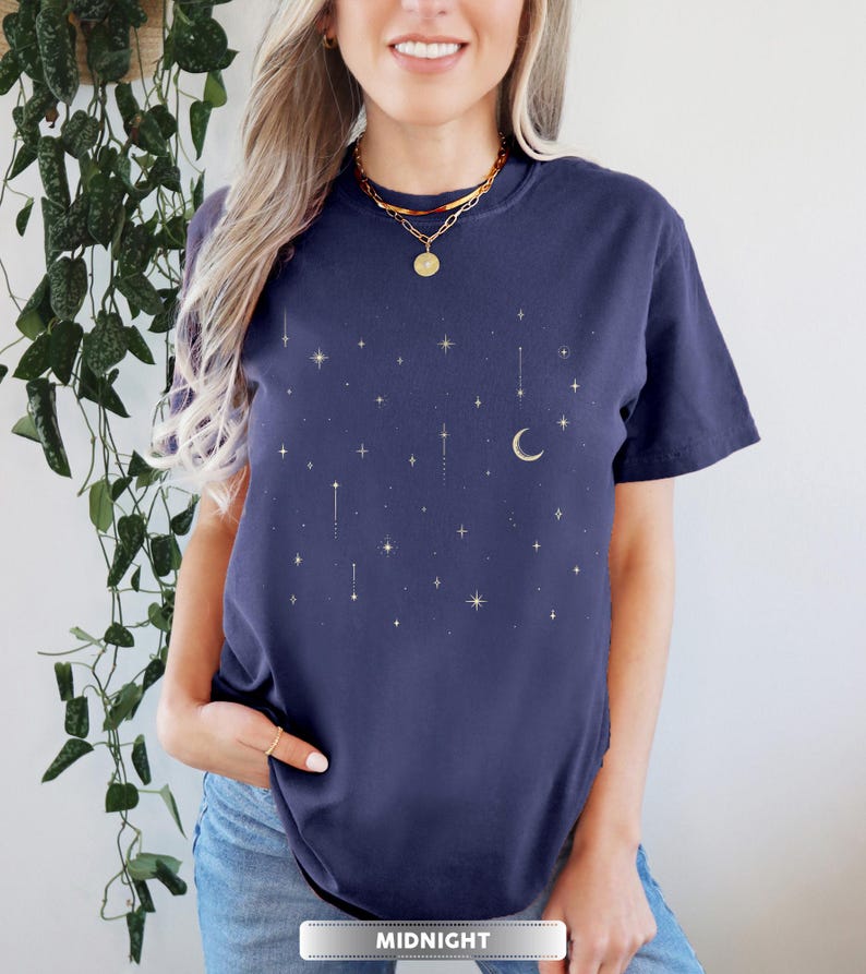May include: Navy blue t-shirt with a celestial design featuring gold stars and a crescent moon. The word "MIDNIGHT" is printed at the bottom. The shirt has a crew neck and short sleeves.
