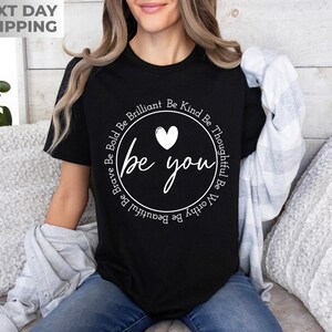 May include: Black t-shirt with a white circle graphic that says "be you" with a heart and the words "Be Kind Be Thoughtful Be Worthy Be Brilliant Be Beautiful Be Brave Be Bold" around the circle.