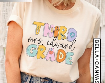 May include: A beige t-shirt with the text "THIRD GRADE" in colorful, outlined letters. The text "mrs. edward" is written in black script below the letters. The letters are decorated with small illustrations of school supplies, such as a pencil, a globe, a paper airplane, and a star.