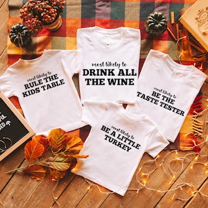May include: White t-shirts with black text, including phrases like "Drink All The Wine" and "Be The Taste Tester." The shirts are arranged on a wooden surface with fall-themed decorations, such as pumpkins and leaves. A letter board reads "Thankful, Grateful, Blessed."