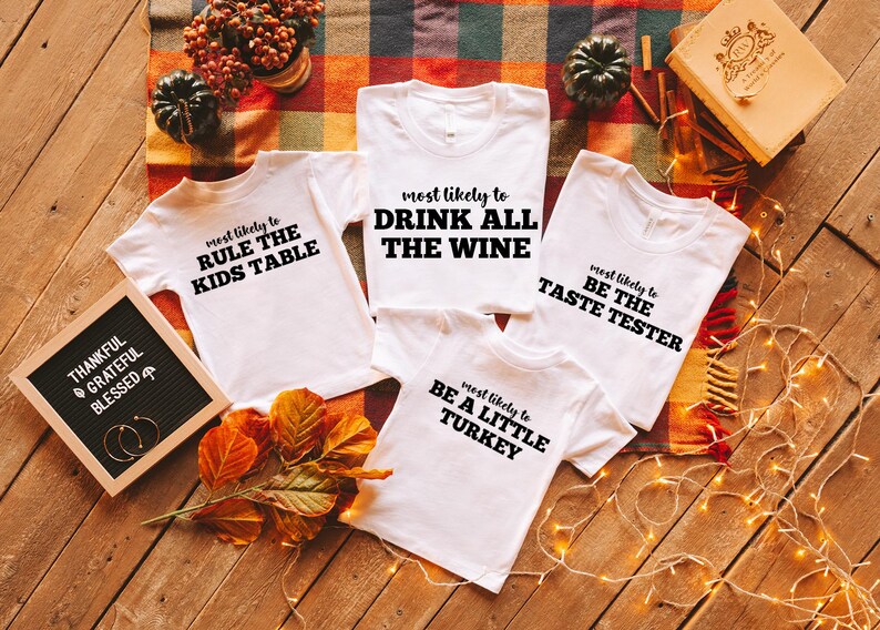 May include: White t-shirts with black text, including phrases like "Drink All The Wine" and "Be The Taste Tester." The shirts are arranged on a wooden surface with fall-themed decorations, such as pumpkins and leaves. A letter board reads "Thankful, Grateful, Blessed."