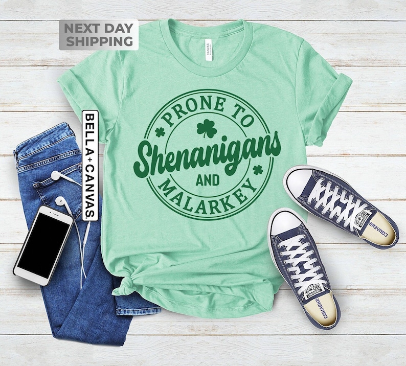 Op de afbeelding: Een lichtgroen T-shirt met een groene cirkelvormige afbeelding met de tekst "Prone to Shenanigans and Malarkey" met een klavertje vier in de cirkel.