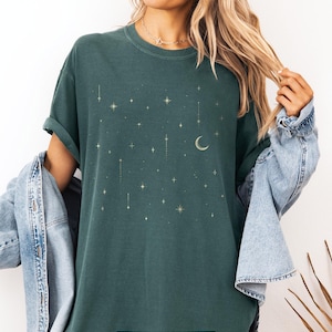 May include: A dark teal t-shirt with a celestial design featuring gold stars and a crescent moon. The shirt has short sleeves and the text "BLUE SPRUCE" is at the bottom. The shirt is part of a clothing collection.