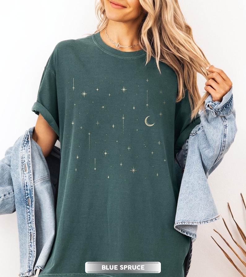 May include: A dark teal t-shirt with a celestial design featuring gold stars and a crescent moon. The shirt has short sleeves and the text "BLUE SPRUCE" is at the bottom. The shirt is part of a clothing collection.