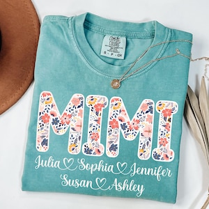 May include: A turquoise Comfort Colors t-shirt with the word "MIMI" in large, floral-patterned letters. Below the word are the names Julia, Sophia, Jennifer, Susan, and Ashley. A gold necklace and a brown hat are also visible.