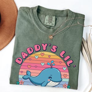 May include: Green t-shirt with a pink and blue graphic of a whale in a sunset with the text "Daddy's Lil'"
