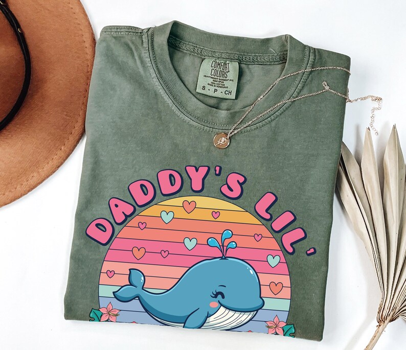 May include: Green t-shirt with a pink and blue graphic of a whale in a sunset with the text "Daddy's Lil'"