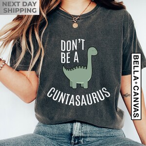 Pu&ograve; includere: Una maglietta grigia scura con il testo "Don't be a Cuntasaurus" in bianco. Un dinosauro verde dei cartoni animati &egrave; raffigurato accanto al testo.