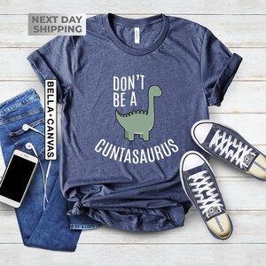 Pu&ograve; includere: Una maglietta blu navy con un'illustrazione di dinosauro verde e il testo "Don't be a Cuntasaurus".