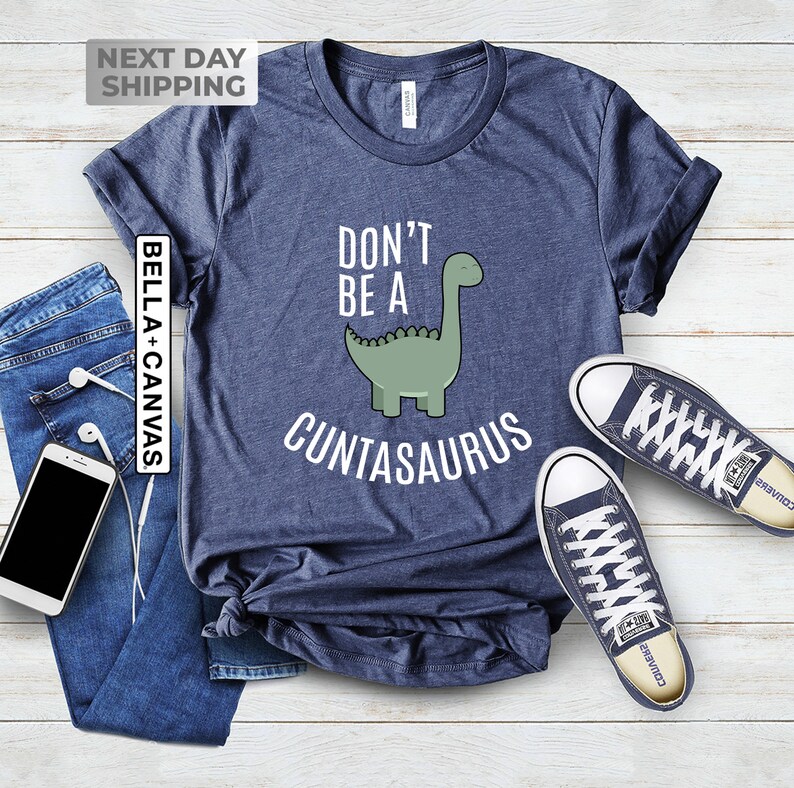 Pu&ograve; includere: Una maglietta blu navy con un'illustrazione di dinosauro verde e il testo "Don't be a Cuntasaurus".