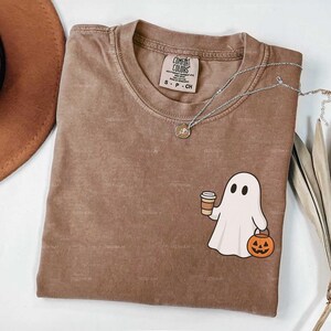 May include: Espresso-coloured Comfort Colors t-shirt with a round neck. The shirt features a white ghost graphic holding a coffee cup and a jack-o'-lantern. A silver necklace with a pendant is draped across the shirt. The shirt is labelled S, P, CH.