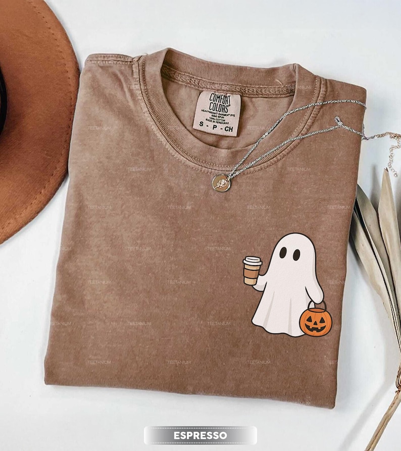 May include: Espresso-coloured Comfort Colors t-shirt with a round neck. The shirt features a white ghost graphic holding a coffee cup and a jack-o'-lantern. A silver necklace with a pendant is draped across the shirt. The shirt is labelled S, P, CH.