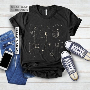 May include: A black t-shirt with a celestial design featuring a crescent moon, stars, and planets. The design is in a cream color. The t-shirt is laid flat on a white wooden surface with a pair of blue converse shoes.