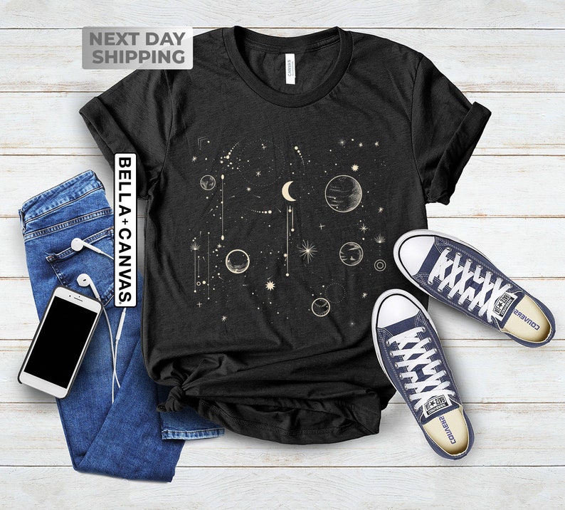 May include: A black t-shirt with a celestial design featuring a crescent moon, stars, and planets. The design is in a cream color. The t-shirt is laid flat on a white wooden surface with a pair of blue converse shoes.