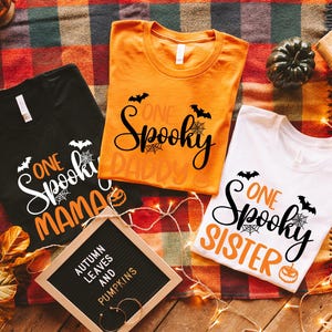 May include: Three Halloween-themed t-shirts in black, orange, and white, each with the words "One Spooky" and a family member title. The shirts feature bats, spiderwebs, and pumpkin graphics. A sign reads "Autumn Leaves and Pumpkins."