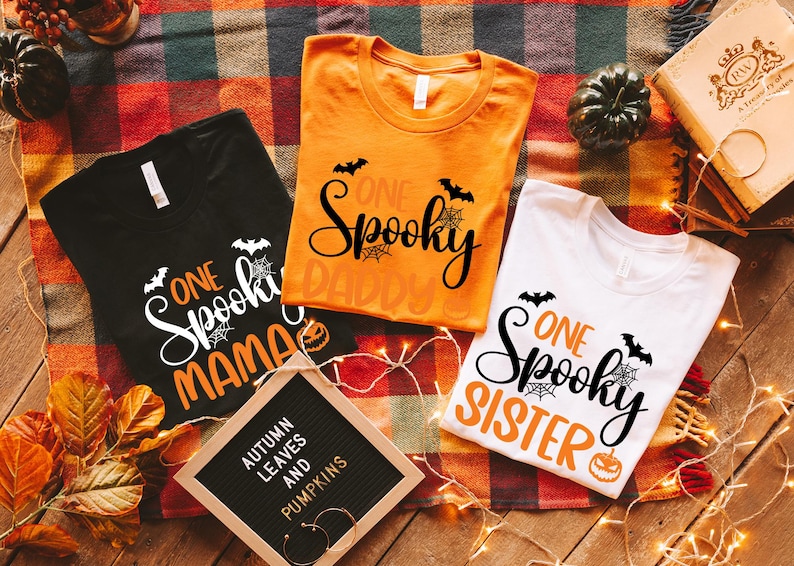 May include: Three Halloween-themed t-shirts in black, orange, and white, each with the words "One Spooky" and a family member title. The shirts feature bats, spiderwebs, and pumpkin graphics. A sign reads "Autumn Leaves and Pumpkins."