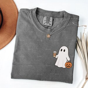 May include: A grey Comfort Colors t-shirt with a round neck, featuring a cartoon ghost holding a coffee cup and a jack-o'-lantern. A silver necklace with a pendant rests on the shirt. The word "PEPPER" is displayed at the bottom.