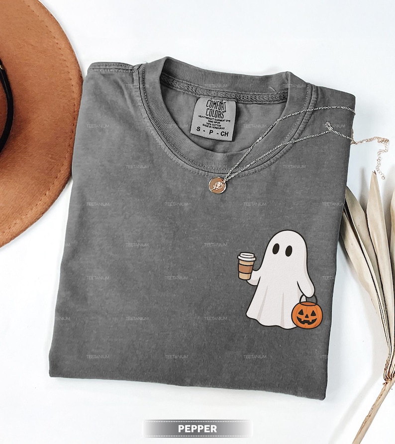 May include: A grey Comfort Colors t-shirt with a round neck, featuring a cartoon ghost holding a coffee cup and a jack-o'-lantern. A silver necklace with a pendant rests on the shirt. The word "PEPPER" is displayed at the bottom.