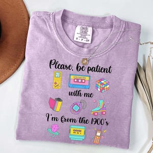 May include: A lilac t-shirt with the text "Please, be patient with me, I'm from the 1900's." The shirt displays colourful graphics of a cassette tape, Rubik's cube, roller skate, and other retro items. A silver necklace is draped across the shirt.