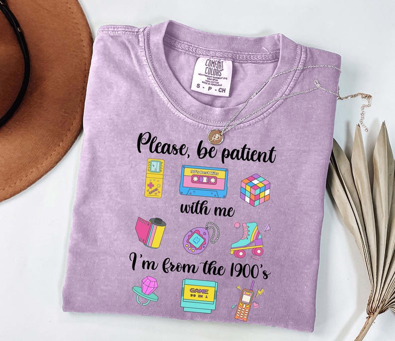 May include: A lilac t-shirt with the text "Please, be patient with me, I'm from the 1900's." The shirt displays colourful graphics of a cassette tape, Rubik's cube, roller skate, and other retro items. A silver necklace is draped across the shirt.